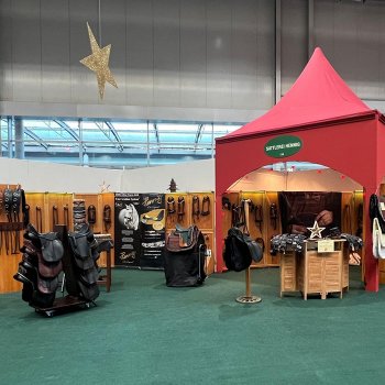 Auf dem Bild sieht man den großen Messestand der Sattlerei Hennig. Man sieht eine Auswahl an Mustersätteln, Zaumzeugen, auch ein Holzpferd mit einem Kindersattel ist im Vordergrund zu sehen. Die Messe ist schön arrangiert, mit roten Bogenpavillons, grünen Teppichen und einem Weihnachtsstern.