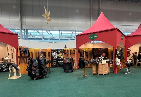Auf dem Bild sieht man den großen Messestand der Sattlerei Hennig. Man sieht eine Auswahl an Mustersätteln, Zaumzeugen, auch ein Holzpferd mit einem Kindersattel ist im Vordergrund zu sehen. Die Messe ist schön arrangiert, mit roten Bogenpavillons, grünen Teppichen und einem Weihnachtsstern.